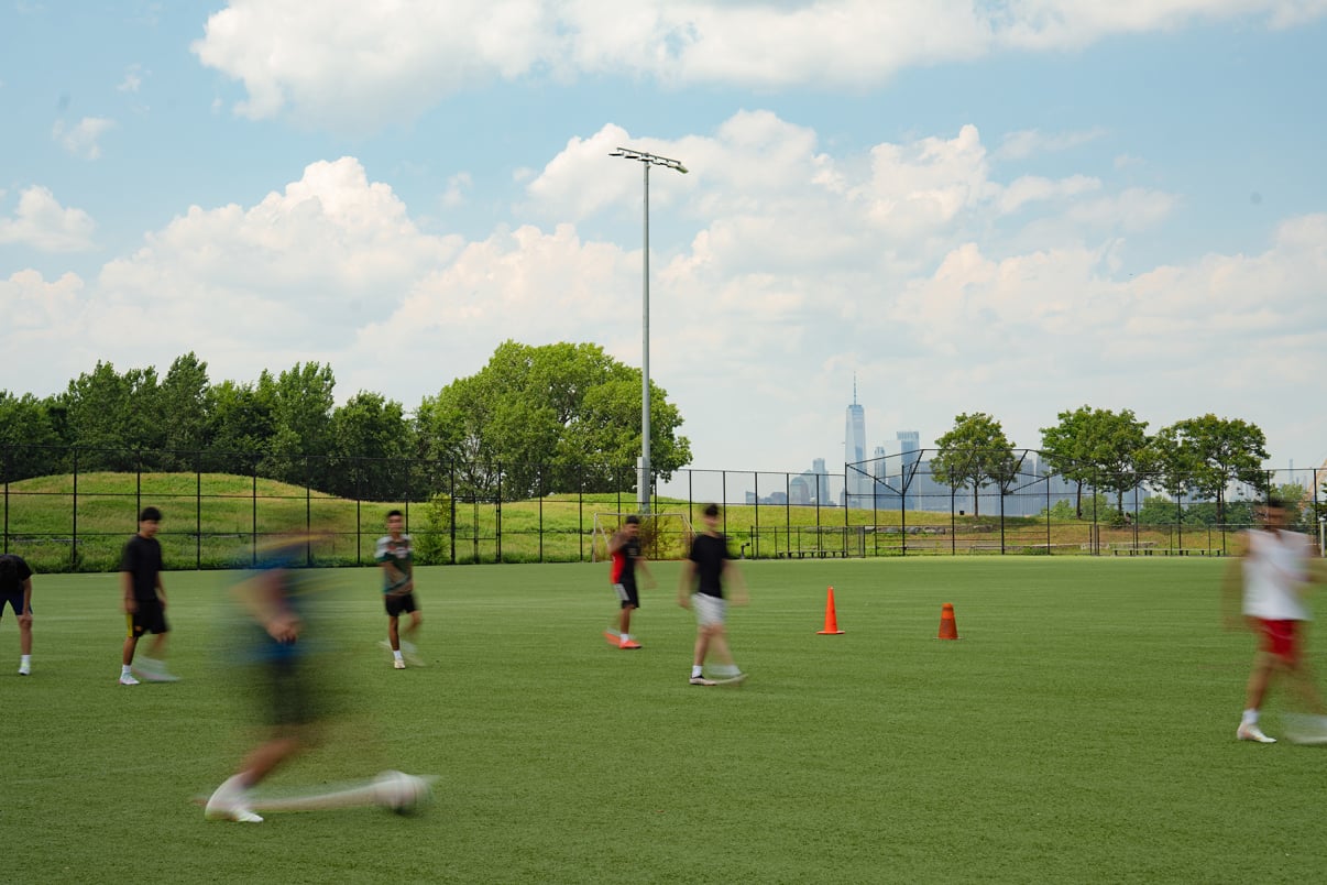 A group of people play soccer on a green field with orange cones, some in motion blur. Trees and a city skyline with tall buildings are visible in the background under a partly cloudy sky.