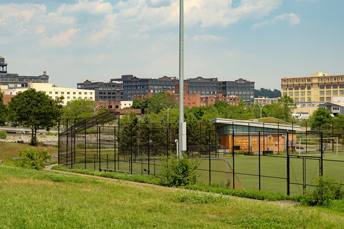 A fenced outdoor sports field with artificial turf is surrounded by greenery and trees, with industrial buildings visible in the background under a partly cloudy sky.