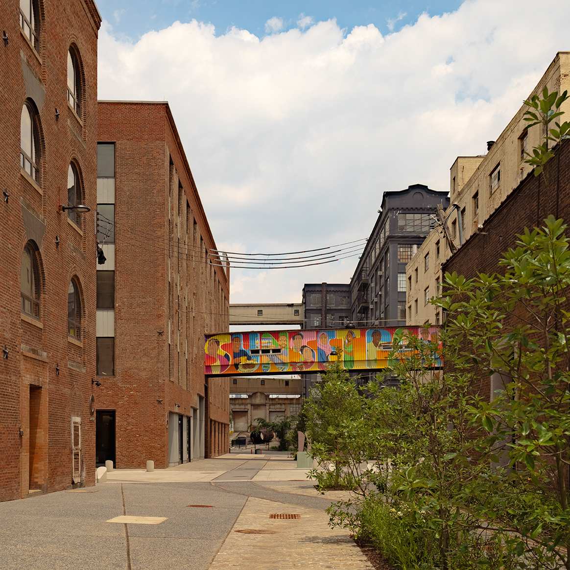 A quiet urban alley lined with brick and concrete buildings, featuring a colorful mural-covered pedestrian bridge connecting two structures, with greenery on the right and a partly cloudy sky above.