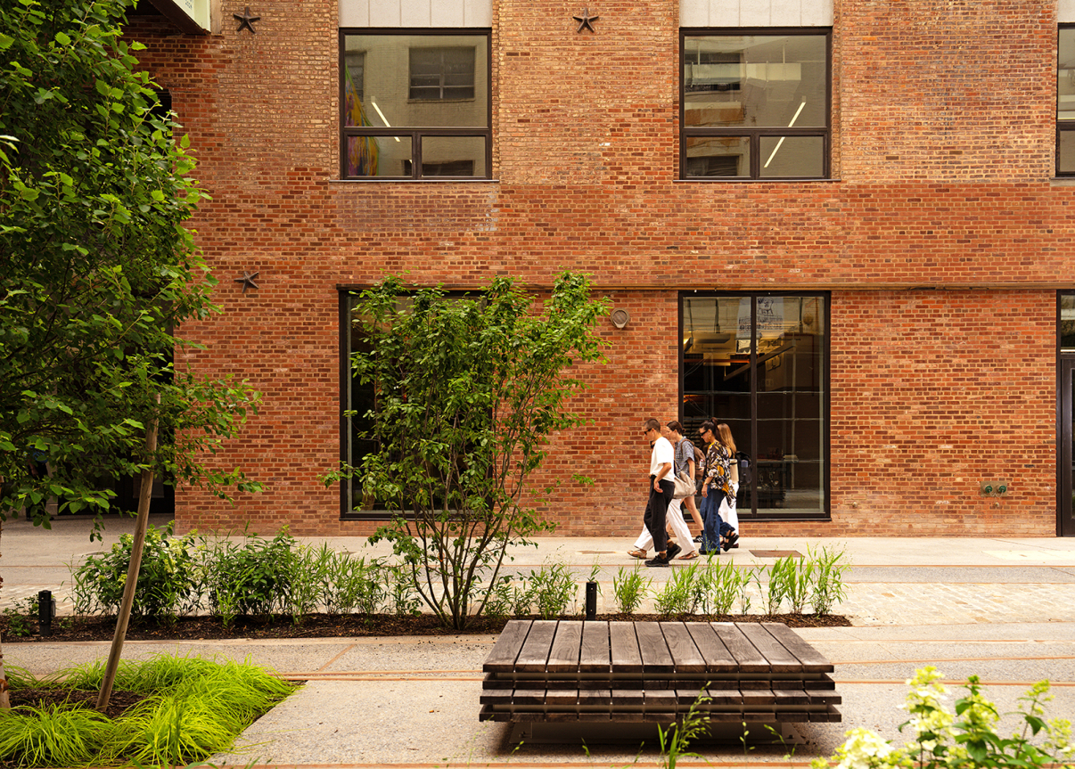Four people walk along a sidewalk next to a brick building with large windows, surrounded by greenery and trees. A wooden bench sits in the foreground of the urban park setting.