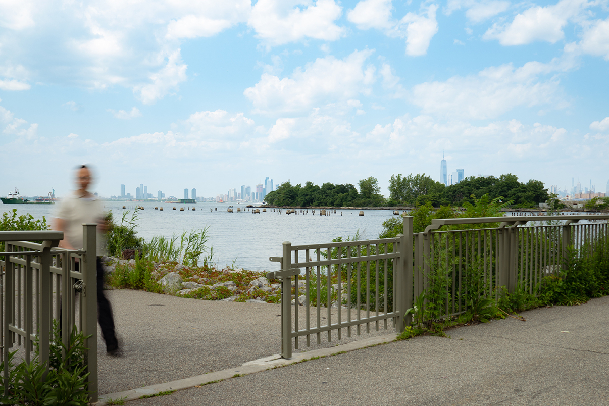 A blurred person walks past a metal fence near a waterfront with distant city skyline and trees under a partly cloudy sky.