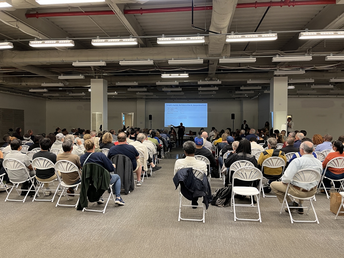 A large group of people sit on folding chairs facing a stage and screen in a spacious indoor venue, attending a presentation or meeting. The room has high ceilings and fluorescent lighting.