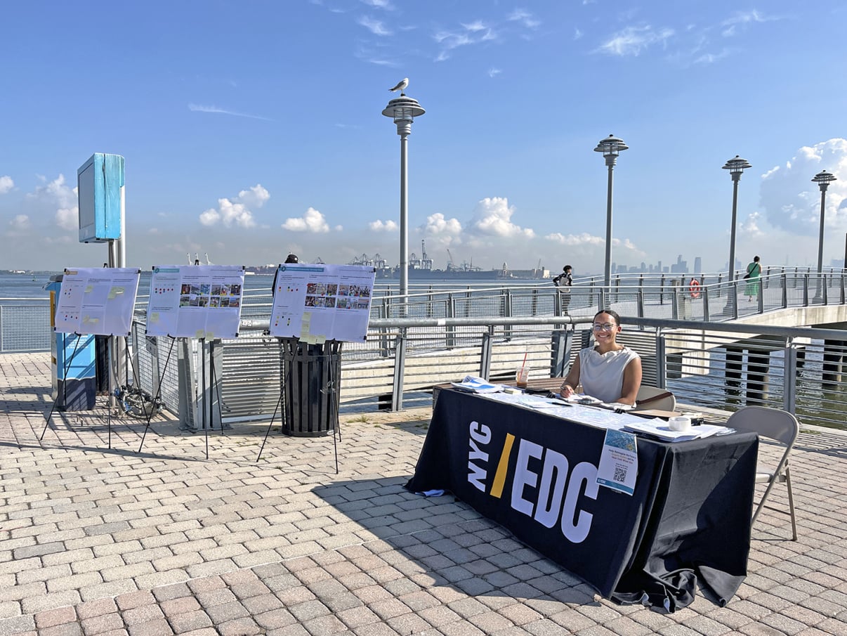 A person sits at an NYC EDC information table on a waterfront promenade with display boards behind them. The sky is blue with scattered clouds, and the city skyline is visible in the distance.