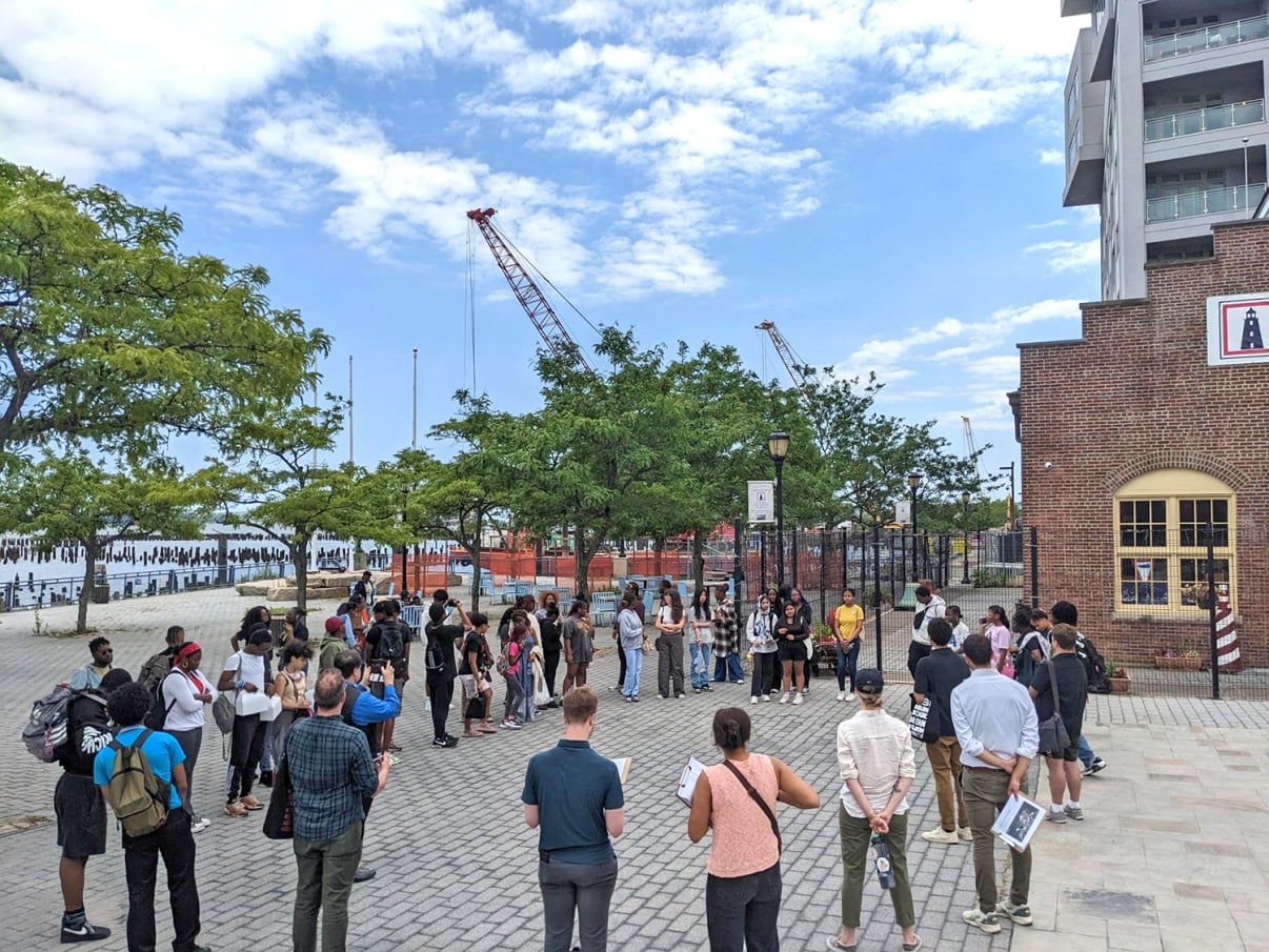 A diverse group of people stands in a semi-circle on a cobblestone plaza near a waterfront, listening to a speaker. There are trees, a brick building, and construction cranes in the background under a partly cloudy sky.