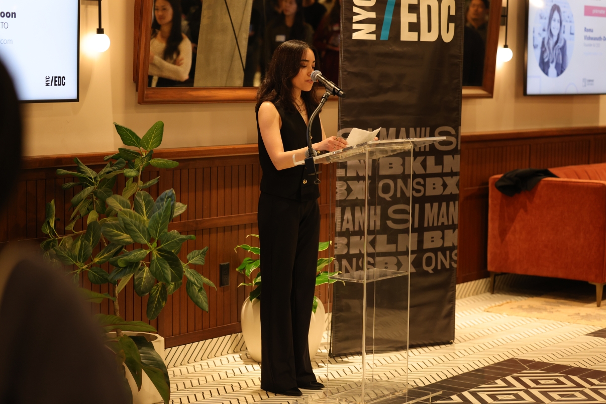 A woman in black speaks at a podium with a microphone at an indoor event. Behind her is a NYCEDC banner, a large plant, and a screen displaying a photo and text. People are reflected in a mirror on the wall.
