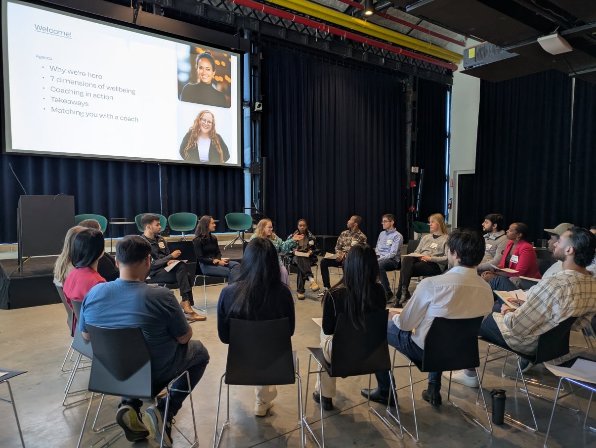 A group of people sit in a circle during a workshop in a modern room. A presentation slide titled Welcome! is displayed on a screen with images of two women and a list of discussion topics.
