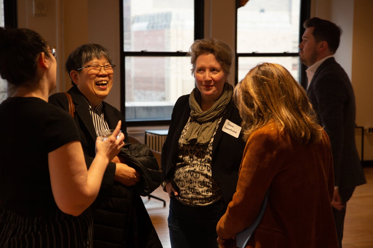 Four adults stand in a warmly lit room, engaged in conversation and smiling at the 2023 Founder Fellowship kickoff. Two women face the camera, one laughing, while others listen. They wear business casual clothes and name tags. Large windows are in the background.