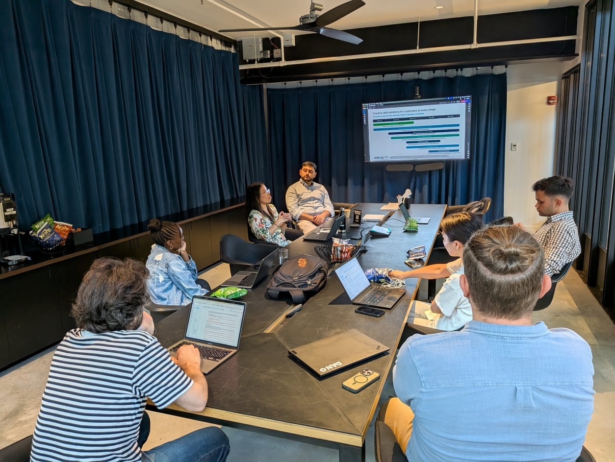 Seven people sit around a conference table, some with laptops, facing a screen displaying a project timeline. The room has dark curtains and modern decor. A mix of men and women are engaged in discussion.
