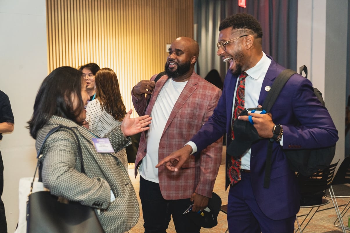 Three people stand together indoors, smiling and laughing as they converse. Two men with backpacks face a woman wearing a patterned coat. Other people are visible in the background near wooden wall panels.