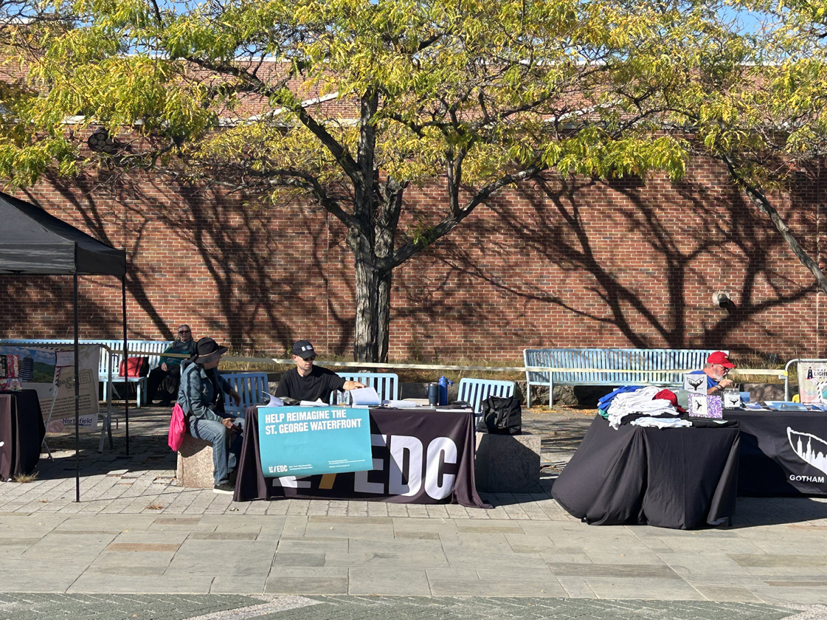 Three outdoor tables are set up under autumn trees, with people sitting behind them. One person holds a large sign about the St. George Waterfront. Blue benches and a brick wall are in the background.