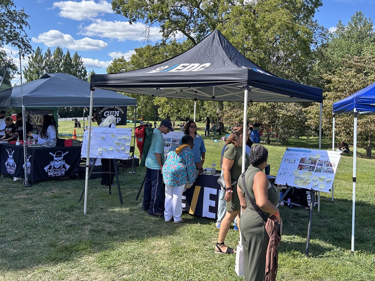 People gather at an outdoor event under black and gray tents on a sunny day in a park. Some read posters and boards with information and sticky notes while others talk. Trees and blue sky with clouds are in the background.