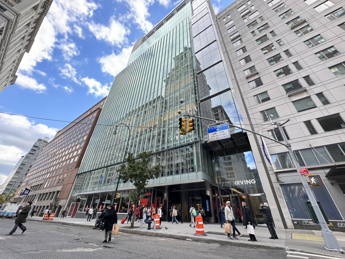 A busy NYC street scene with pedestrians walking near a modern glass building, traffic cones, and tall buildings on either side under a partly cloudy sky.