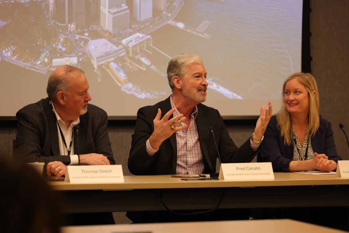 Three panelists sit at a conference table. The man in the center, Fred Cerullo, gestures with his hands while talking, and the others listen to him. A cityscape image is displayed on a projector screen behind them. Name placards are on the table.