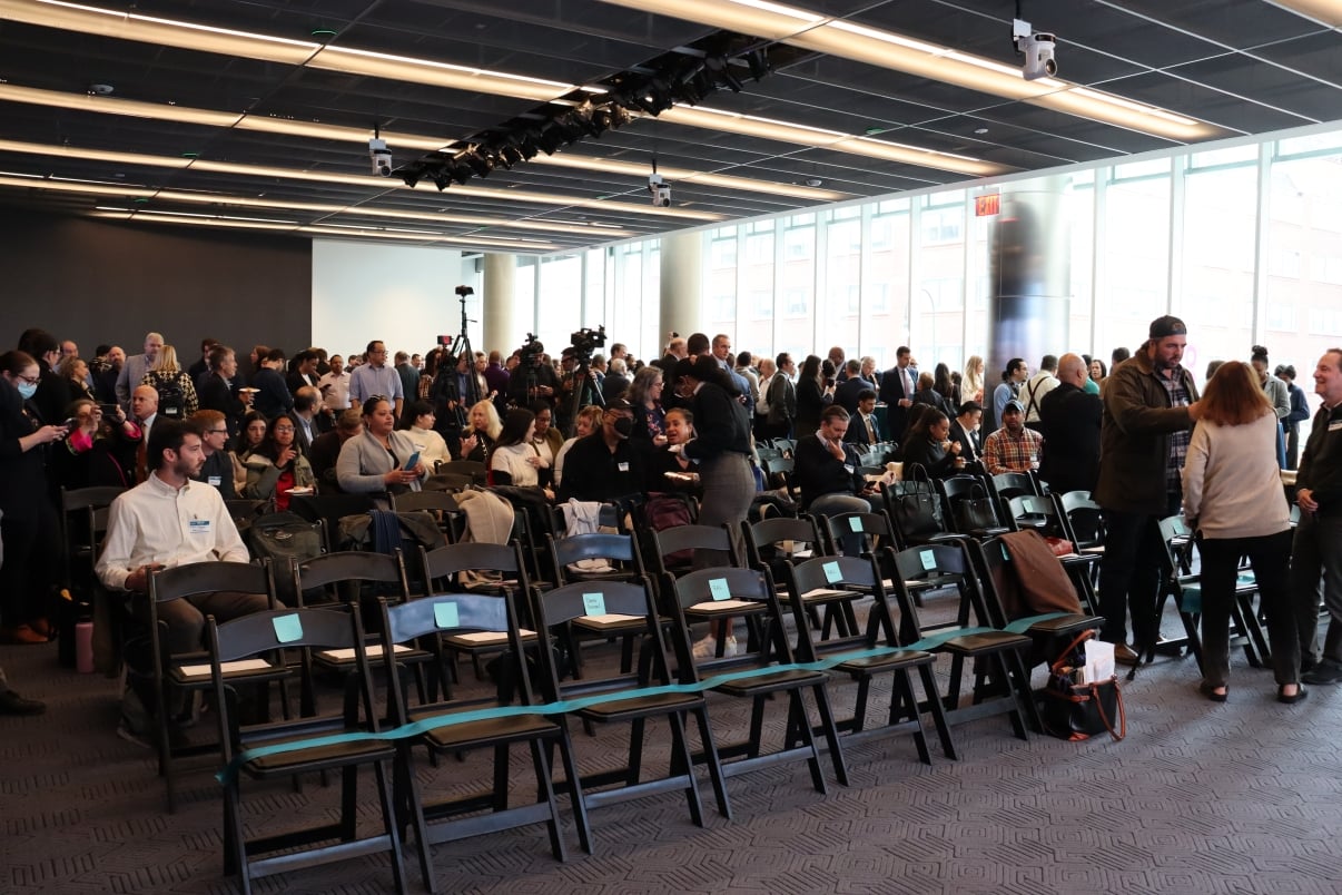 A large group of people gathers in Civic Hall, a modern conference room, with rows of empty chairs in the foreground and bright windows along one wall. Some people are standing, talking, or preparing for the Choose NYC 2025 Summit event to begin.