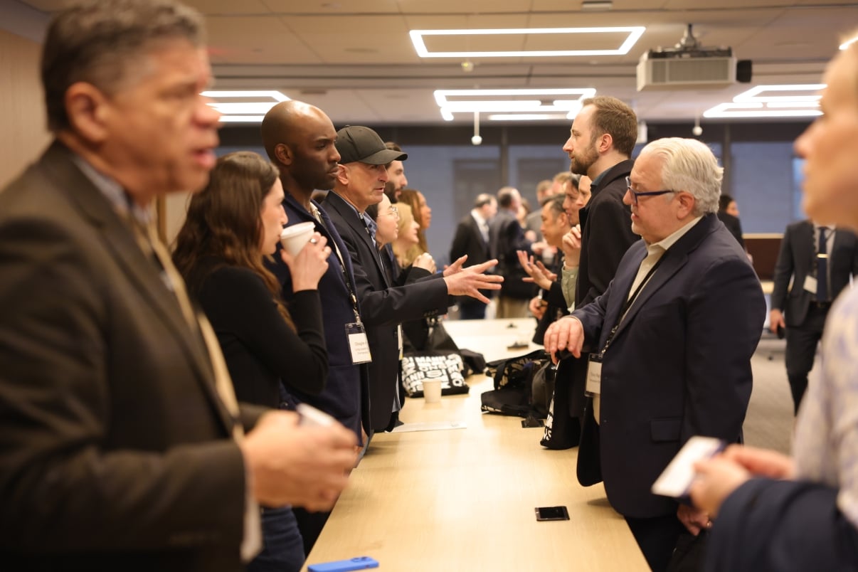 People in business attire stand on both sides of a long table, engaging in animated discussions and networking at the Choose NYC 2025 Summit in a modern NYCEDC conference room.