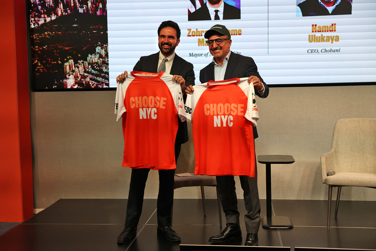 Mayor Mamdani and Hamdi Ulukaya, CEO of Chobani stand on stage holding red and white sports jerseys that say CHOOSE NYC. They are smiling, and a projection screen with names and titles is visible behind them.