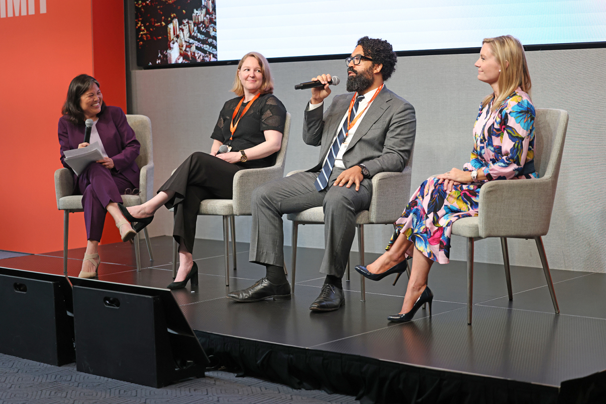 Four people sit on a stage panel; one man speaks into a microphone while the other three, all women, listen and smile. They wear business attire and have event lanyards. An orange backdrop is visible on the left.