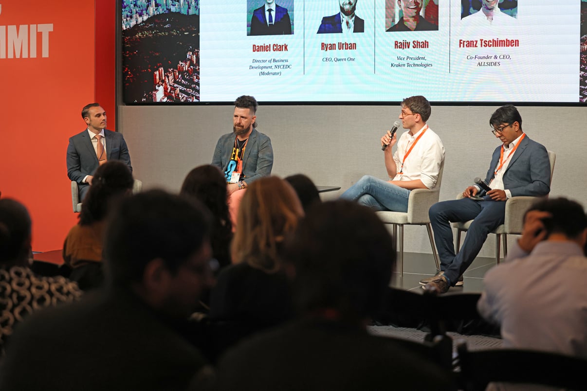 A panel of four men sits on stage at a business summit, three on chairs and one on a stool, with a projected screen behind them displaying names, titles, and photos. Audience members are seated in the foreground.
