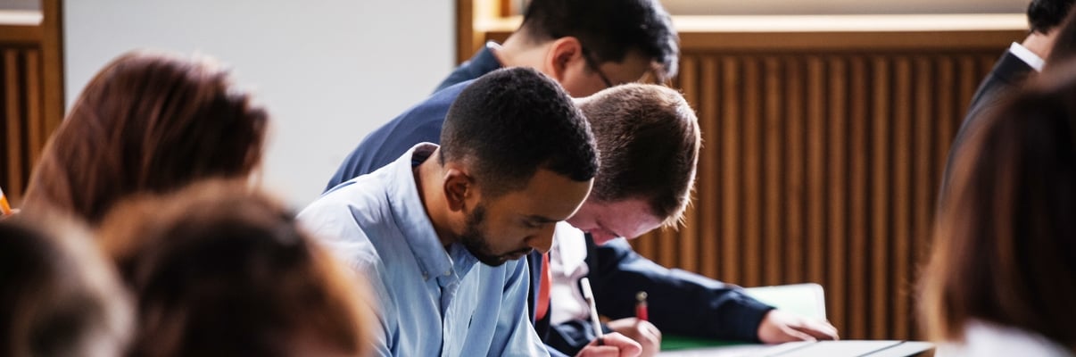 A group of LifeSci NYC applicants sitting at desks in a classroom, focused on writing in notebooks. The students are diverse, dressed in casual attire, and are positioned in rows. The setting is well-lit, with wooden paneling on the walls. Photo by Jordan Rathkopf/NYCEDC