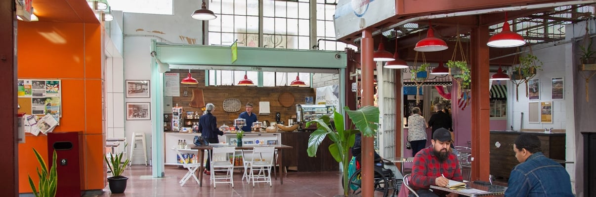 Indoor view of La Marqueta Market with a few people seated at tables. The space has high ceilings, red and orange walls, and greenery. There are various food stalls, and a large window allows natural light to fill the area. Indoor view of La Marqueta Market with a few people seated at tables. The space has high ceilings, red and orange walls, and greenery. There are various food stalls, and a large window allows natural light to fill the area.