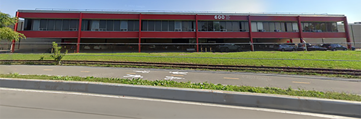 Exterior view of 600 Food Center Drive. A modern two-story red building with numerous windows, set against a clear blue sky with scattered clouds. A road and railroad tracks run parallel to it in the foreground. A few cars are parked beside the building.