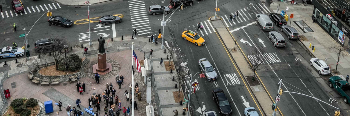 Aerial view of Chatham/Kimlau Square in New York City&#039;s Chinatown. A busy intersection with many cars, taxis, and people crossing the street. A small park with a statue and American flags is visible in the lower left corner. Multiple crosswalks and traffic signals are present.