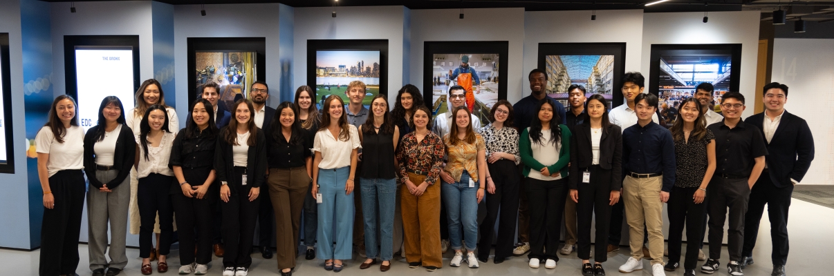 A group of 28 NYCEDC Summer 2024 Interns stand together at the reception area on the 14th floor, posing for a group photo in front of digital screens displaying various images.