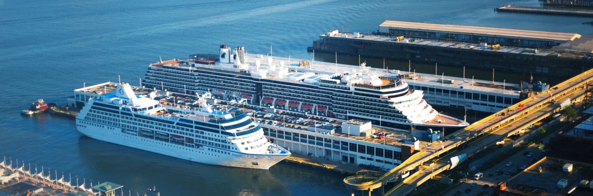 Aerial view of Manhattan cruise ships docked at Hudson River piers in Manhattan Cruise Terminal, with buildings and a waterfront in the background.