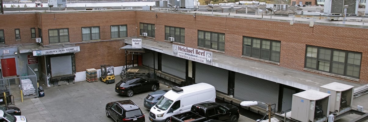 Aerial view of Gansevoort Square: A brick industrial building with loading docks, parked cars, and a sign reading "Mitchell Beef." Urban landscape visible in the background. Aerial view of Gansevoort Square: A brick industrial building with loading docks, parked cars, and a sign reading "Mitchell Beef." Urban landscape visible in the background.