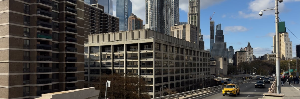 Urban scene at 100 Gold Street with a yellow taxi driving on a bridge in Manhattan. Tall skyscrapers are in the background beneath a partly cloudy sky. The setting appears busy and modern with multiple lanes on the road.