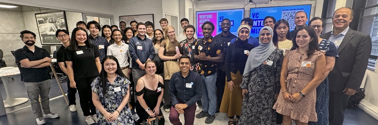 A diverse group of people, some standing and some kneeling, smile for a group photo in an indoor setting. They are at an event for the NYC Venture Capital Internship Program, with name tags visible on their clothing. A screen in the background adds context to the meeting.