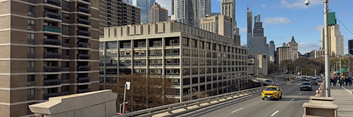 Urban scene at 100 Gold Street with a yellow taxi driving on a bridge in Manhattan. Tall skyscrapers are in the background beneath a partly cloudy sky. The setting appears busy and modern with multiple lanes on the road.