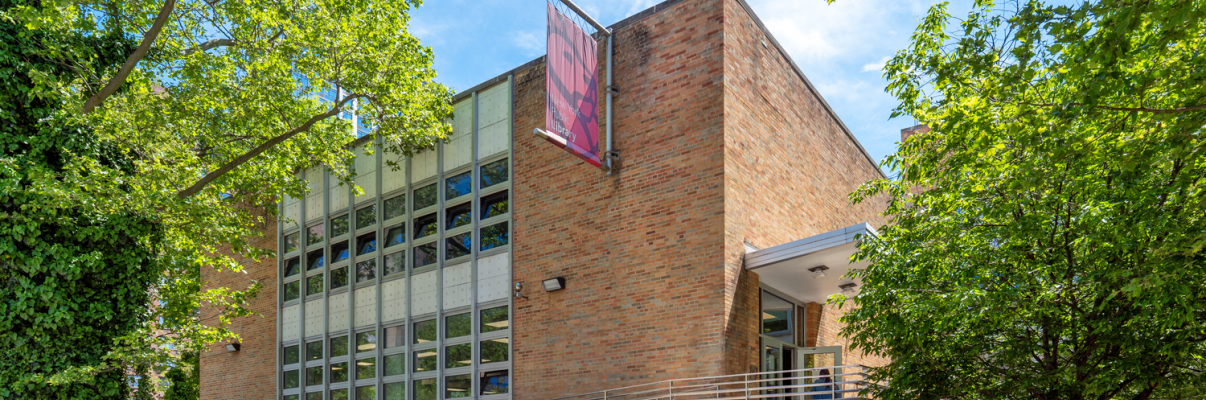 A modern brick building with large grid-like windows, a sidewalk with a metal fence in front, and leafy green trees surrounding the entrance. A red New York Public Library banner hangs near the entrance on a sunny day. A modern brick building with large grid-like windows, a sidewalk with a metal fence in front, and leafy green trees surrounding the entrance. A red New York Public Library banner hangs near the entrance on a sunny day.