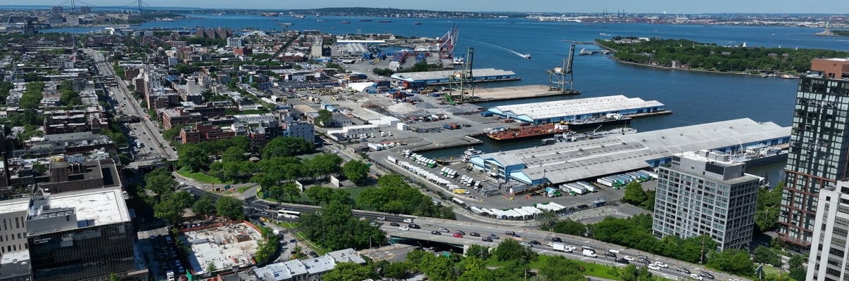 Aerial view of the Brooklyn Marine Terminal with industrial buildings, shipping docks, highways, green spaces, and a river; bridges and islands are visible in the distance under a clear blue sky. Aerial view of the Brooklyn Marine Terminal with industrial buildings, shipping docks, highways, green spaces, and a river; bridges and islands are visible in the distance under a clear blue sky.