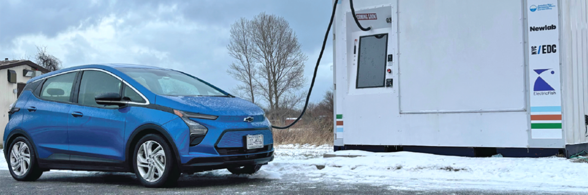 A blue electric car is parked on a snowy roadside next to a white charging station. The sky is overcast, and leafless trees are visible in the background. The charging station features logos and a cable plugged into the car.