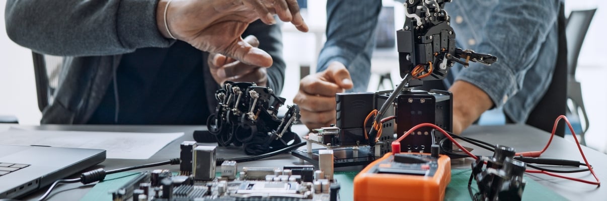Two people work together on assembling and adjusting a robotic hand at a desk covered with electronic components, wires, and circuit boards.