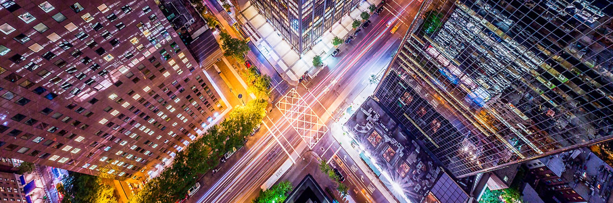 A vibrant, aerial night view of a New York City intersection surrounded by tall skyscrapers, with colorful lights from buildings and streaks of headlights and taillights showing busy street traffic below.