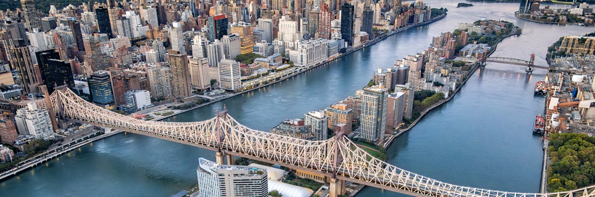 Aerial view of Roosevelt Island and the Ed Koch Queensboro Bridge spanning the East River, connecting Manhattan and Queens in New York City, with tall buildings and lush green spaces along the waterfront.