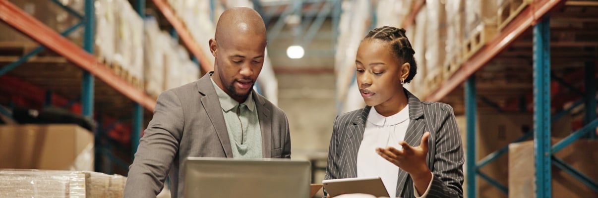 Two people in business attire stand in a warehouse, looking at a laptop and tablet on a table with safety helmets. They appear to be discussing work among shelves of boxes and goods in the background.