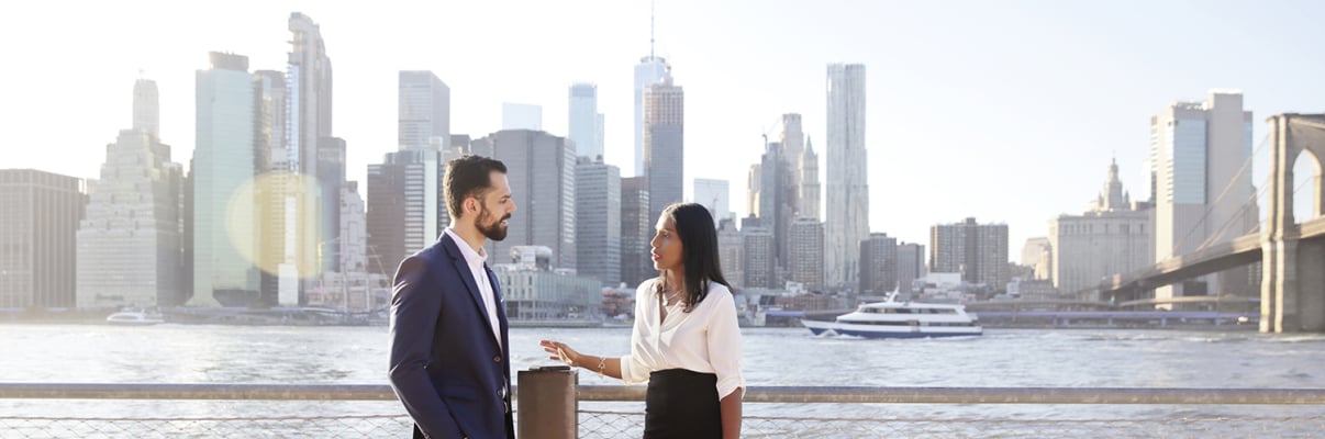 Two professionals in business attire, holding briefcases, have a conversation in front of the New York City skyline.