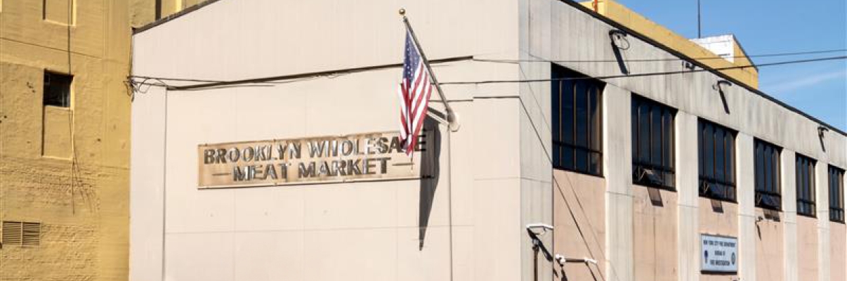 A beige two-story building with an American flag and a sign reading “Brooklyn Wholesale Meat Market” on the front, located on a street corner with yellow-painted curbs.