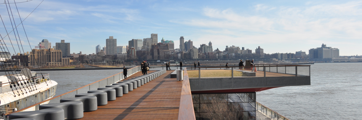 Photo of East River Waterfront Esplanade shows a wooden boardwalk with circular black seats overlooking a river. People are walking along the path, with a ship on the left and a New York City skyline in the distance under a partly cloudy sky.