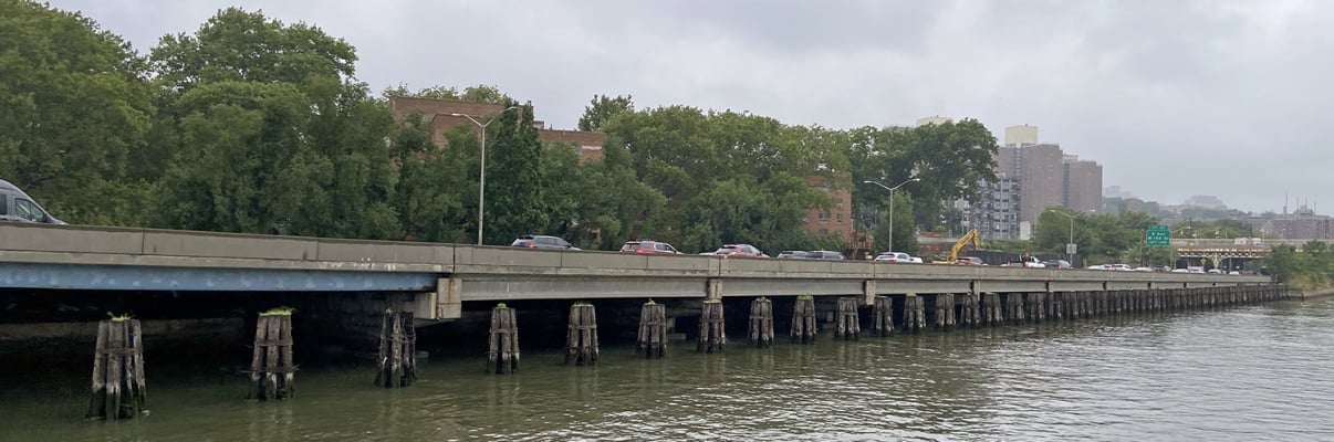 A gray, cloudy day over a riverside highway with cars driving along it. The highway is supported by wooden pilings, with trees and brick buildings visible in the background, and water in the foreground.