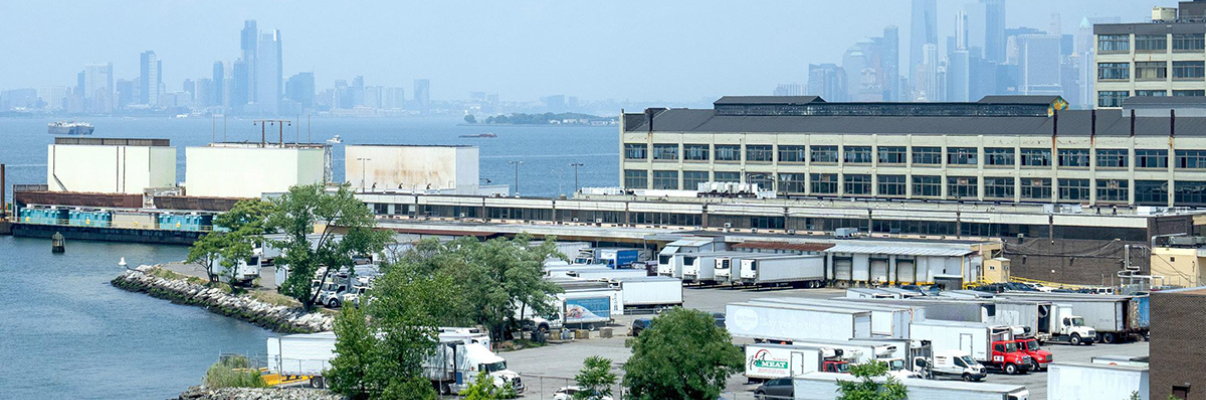 A view of a waterfront industrial area with warehouses, trucks, and greenery; a body of water is in the foreground, and a hazy city skyline is visible in the distance.
