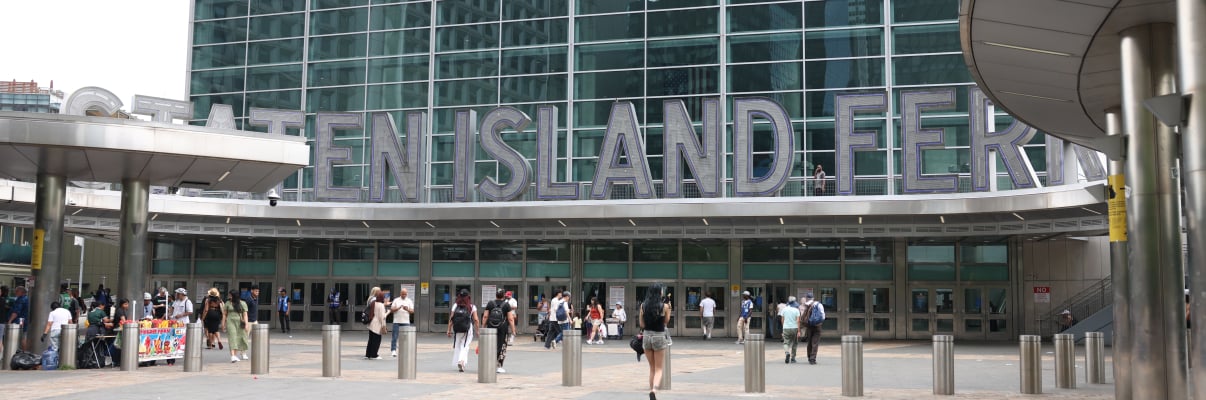 People walk in front of the Staten Island Ferry terminal, a large glass building with “Staten Island Ferry” in big silver letters above the entrance. Some street vendors are set up near the front.