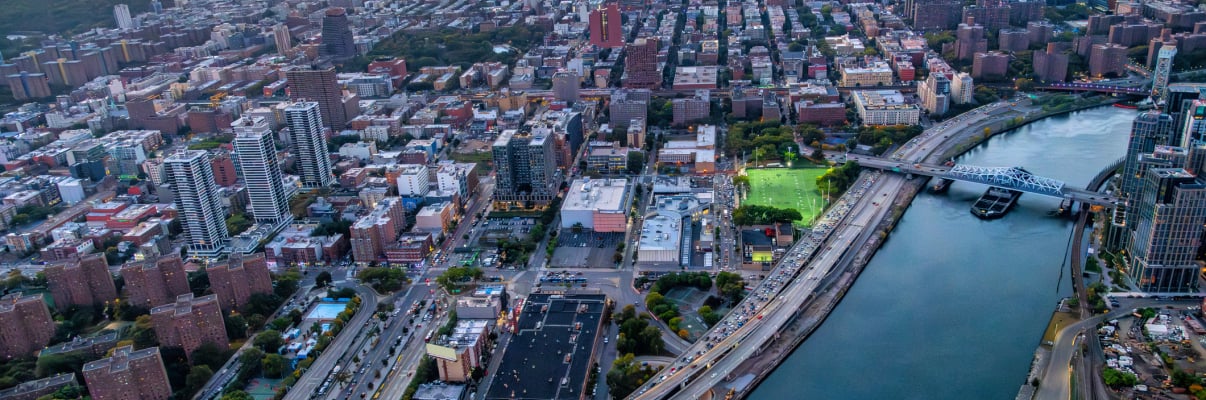 Aerial view of New York City at sunset, showing a river, several bridges, high-rise buildings, busy roads, and a park with green fields. The sky is orange and blue as the sun sets in the distance.