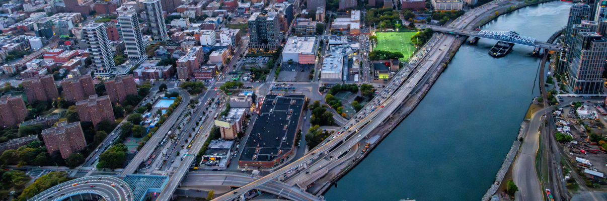 Aerial view of New York City at sunset, showing a river, several bridges, high-rise buildings, busy roads, and a park with green fields. The sky is orange and blue as the sun sets in the distance.