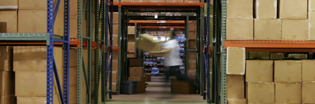A person in motion, carrying boxes in a warehouse aisle filled with stacked brown cardboard boxes on metal shelves. The floor is concrete, and the shelves extend high, suggesting a large storage facility.